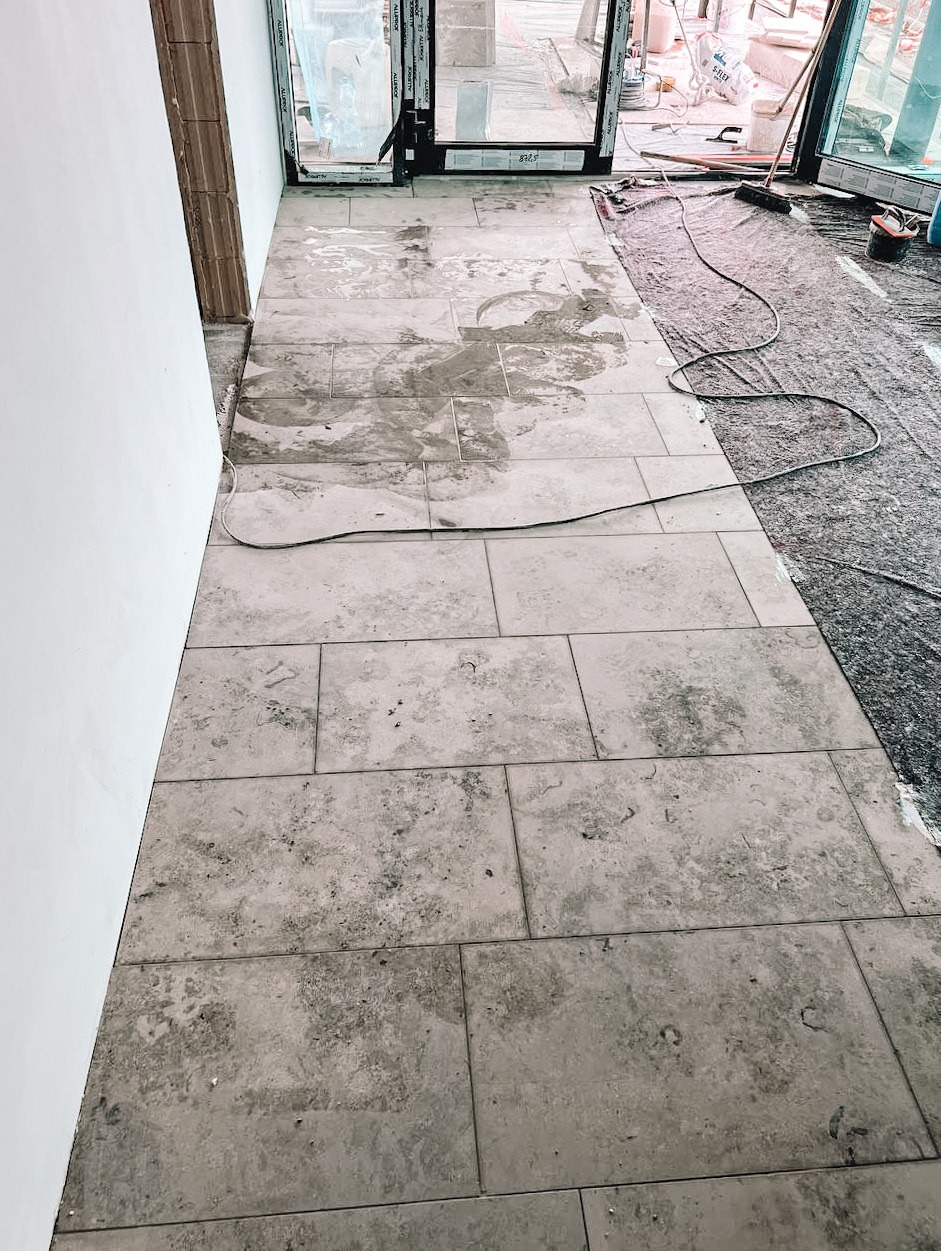 Układanie jasnych, prostokątnych płytek podłogowych z efektem kamienia, częściowo mokrych, w trakcie prac wykończeniowych przy drzwiach balkonowych.