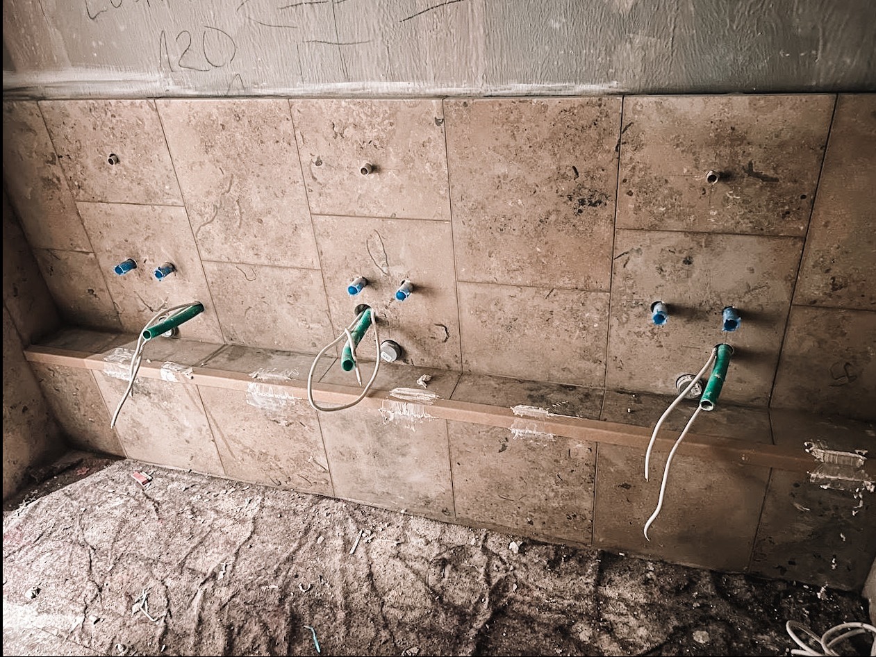 Tarasowa mozaika ścienna w łazience, widoczne wystające rury i kable, przygotowane do montażu armatury. Surowy stan wykończenia, ślady zaprawy.