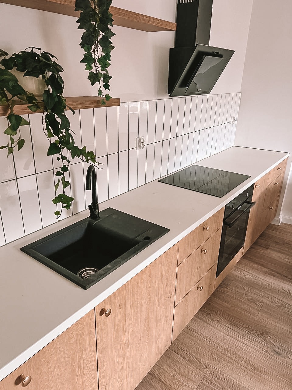 Jasna kuchnia po remoncie: drewniane szafki, biały blat, czarny zlew i płyta indukcyjna. Nad blatem białe, wąskie płytki i czarny okap. Roślina w doniczce.