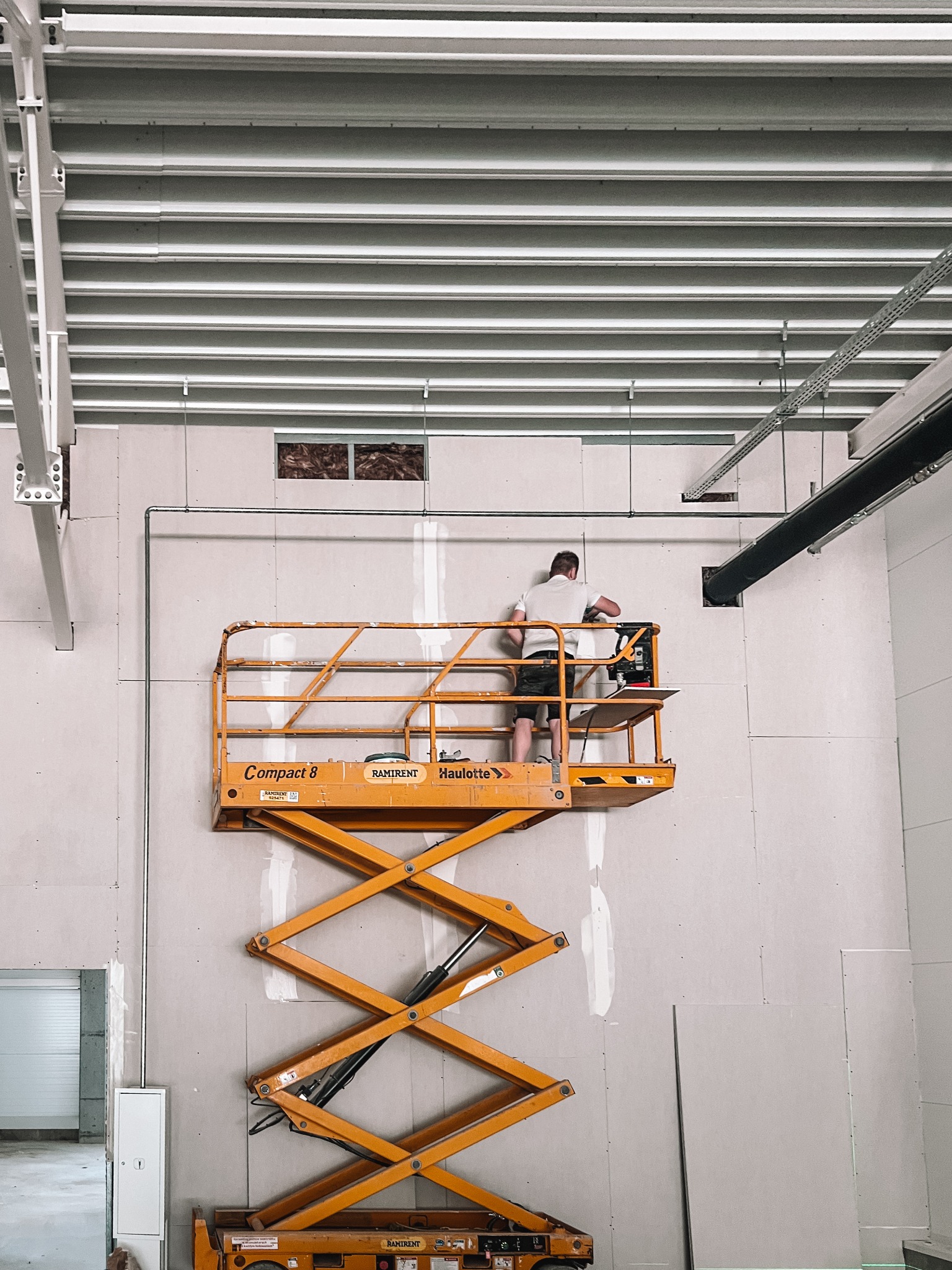 Praca na podnośniku: montaż płyt gipsowych na ścianie w surowym wnętrzu. Widoczne instalacje, oświetlenie sufitowe i fragment otwartego pomieszczenia w tle.