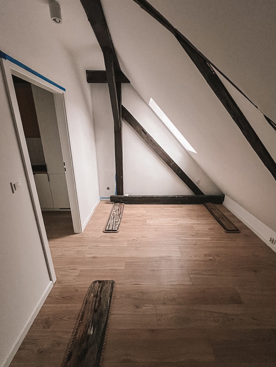 Pokój na poddaszu po remoncie: drewniana podłoga, białe ściany, widoczne elementy konstrukcyjne. Skosy i okno dachowe. Listwy przypodłogowe w trakcie montażu.