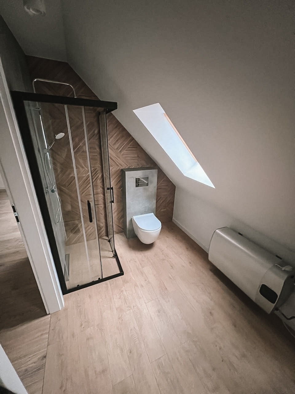 Nowoczesna łazienka na poddaszu ze skośnym oknem dachowym. Kabina prysznicowa z czarną ramą, drewnopodobne płytki i podwieszana toaleta.