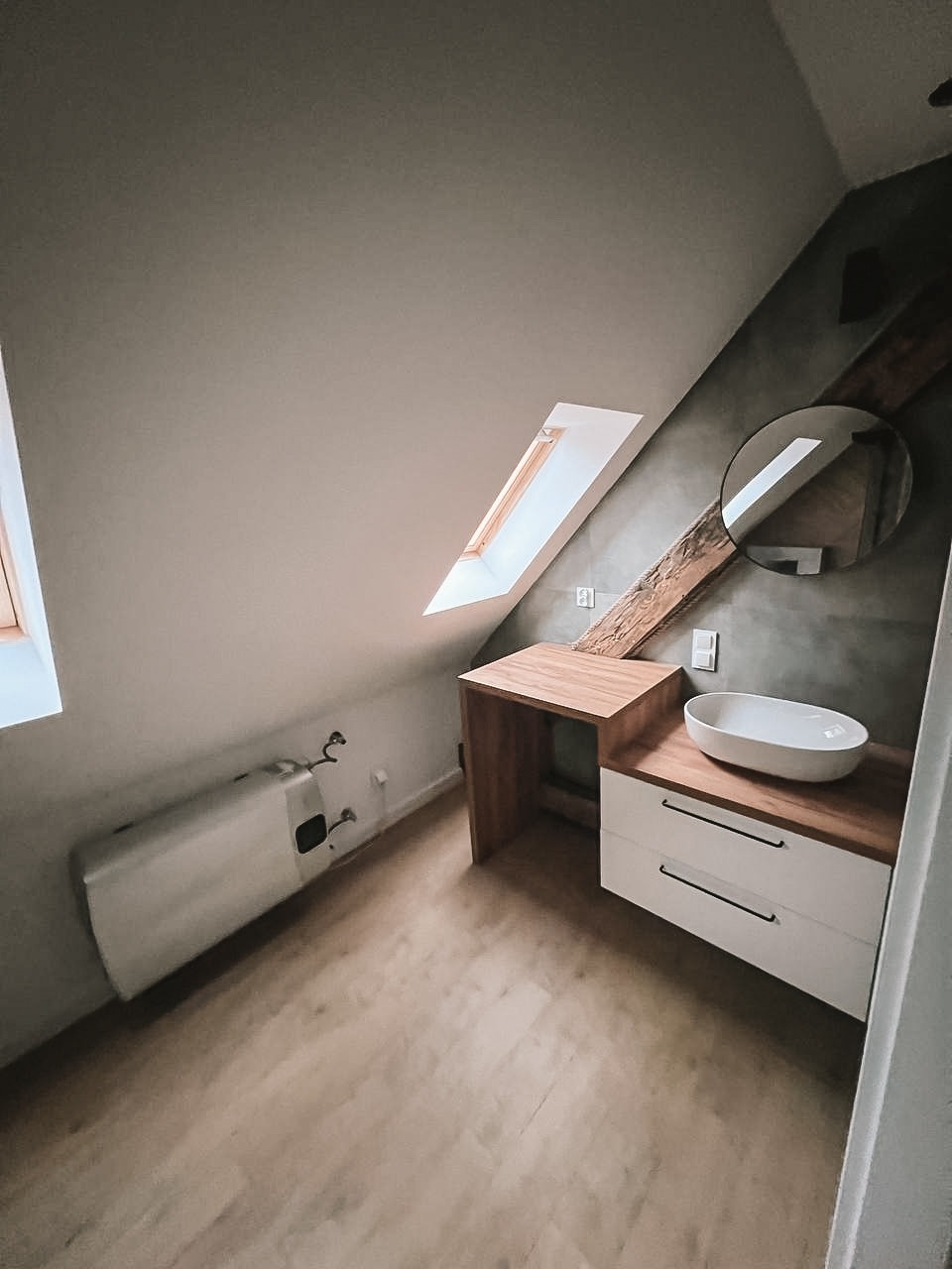 Nowoczesna łazienka na poddaszu z drewnianymi akcentami: umywalka nablatowa, białe szafki, okrągłe lustro i okno dachowe. Minimalistyczny design, szara ściana.
