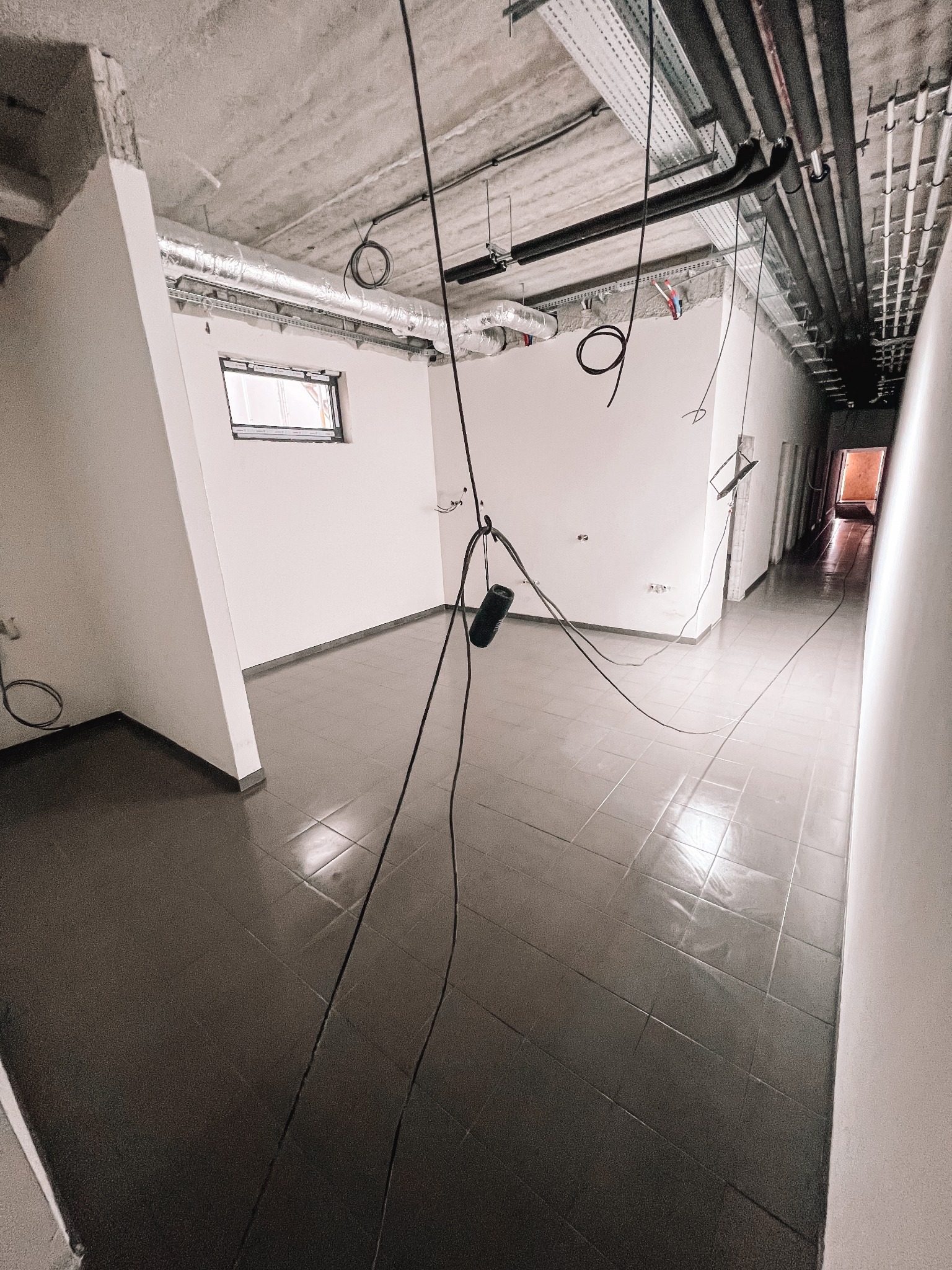 Wnętrze w trakcie prac wykończeniowych: widoczne przewody, rury pod sufitem i szare płytki na podłodze. Małe okno w oddali, surowy betonowy sufit.