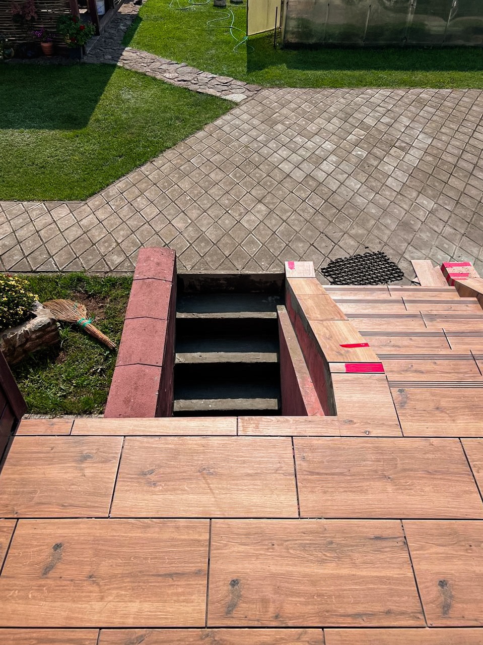 Wykończenie schodów zewnętrznych płytkami imitującymi drewno, prowadzących do piwnicy. Widoczne fragmenty trawnika i kostki brukowej. Czerwone akcenty na krawędziach schodów.