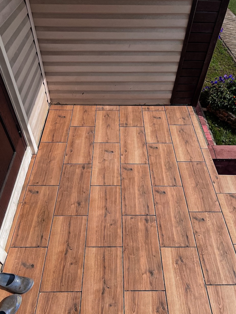 Balkon wyłożony płytkami imitującymi drewno, z widocznym fragmentem elewacji i butami w rogu. Płytki ułożone równo, tworzą estetyczne wykończenie.