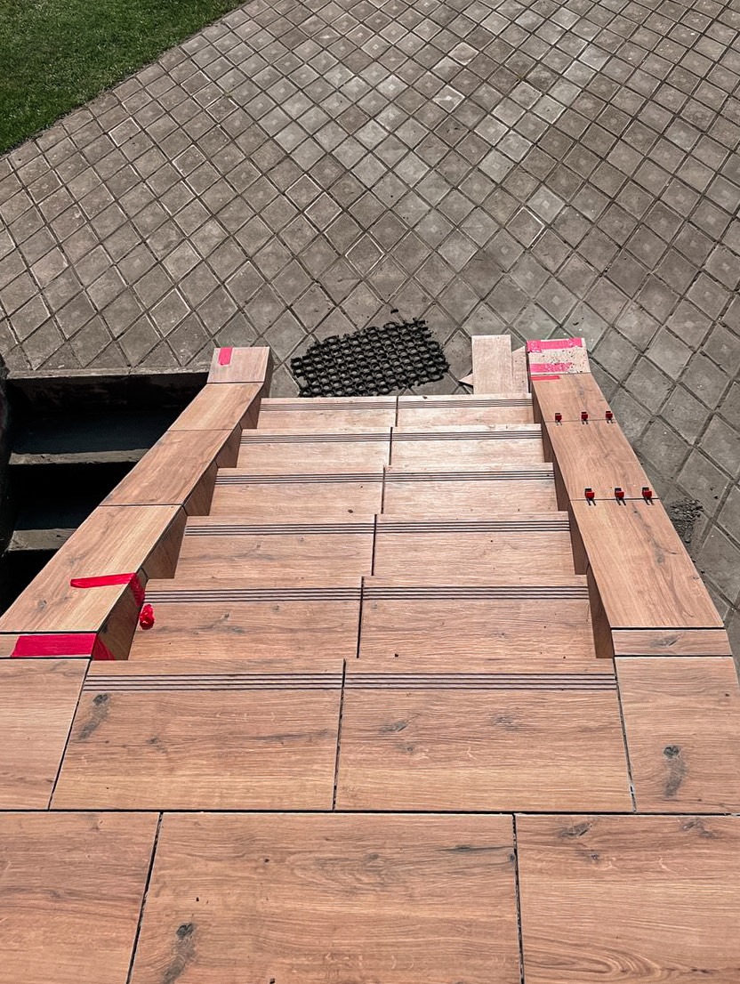 Schody wyłożone płytkami imitującymi drewno we Wrocławiu, widok z góry. Czerwone elementy mocujące widoczne na stopniach. Na górze schodów mata antypoślizgowa.