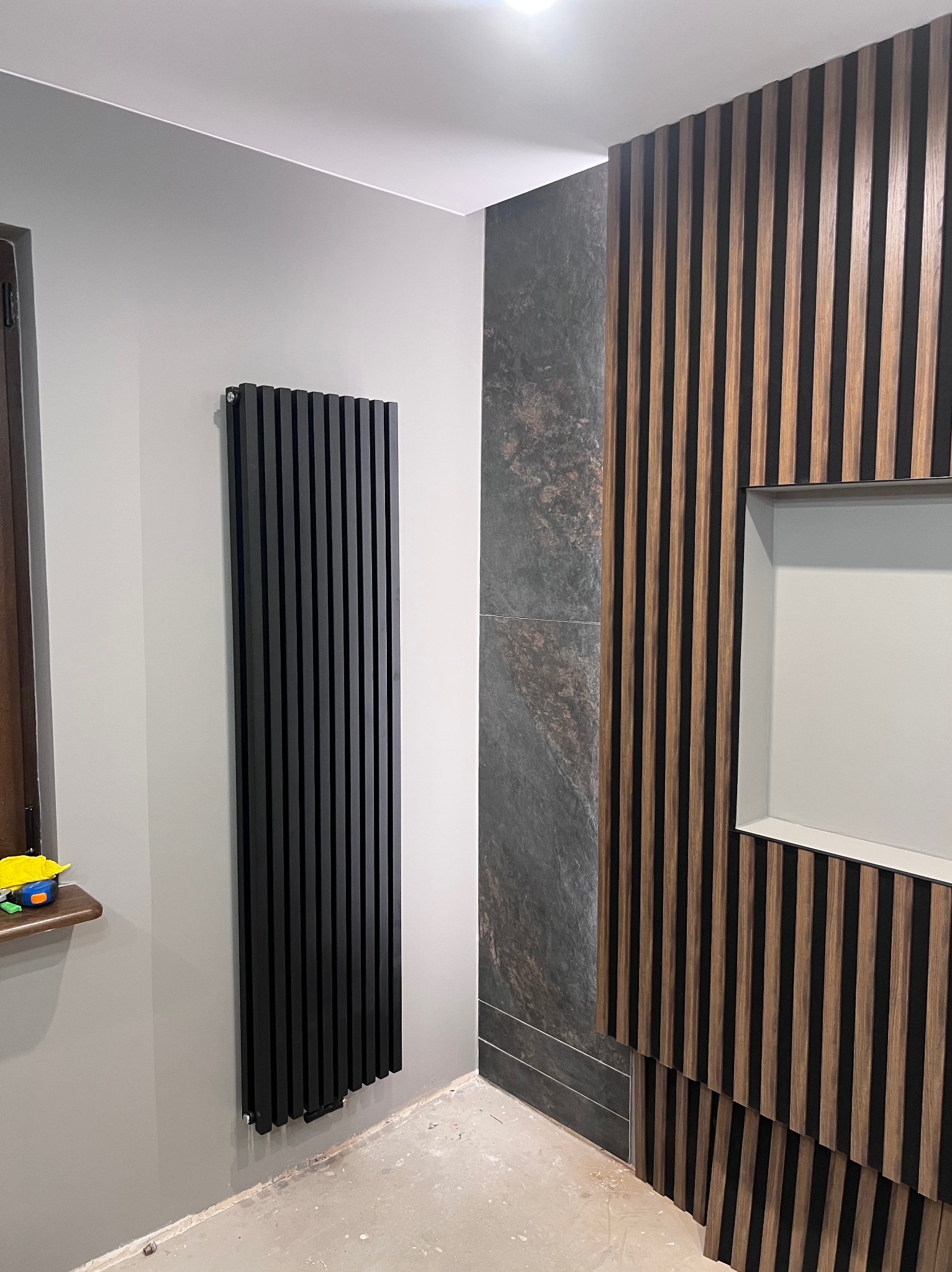Nowoczesna aranżacja ściany z drewnianymi lamelami i płytą imitującą kamień. Czarny, pionowy grzejnik na szarej ścianie. Widok na fragment okna z zabawką.