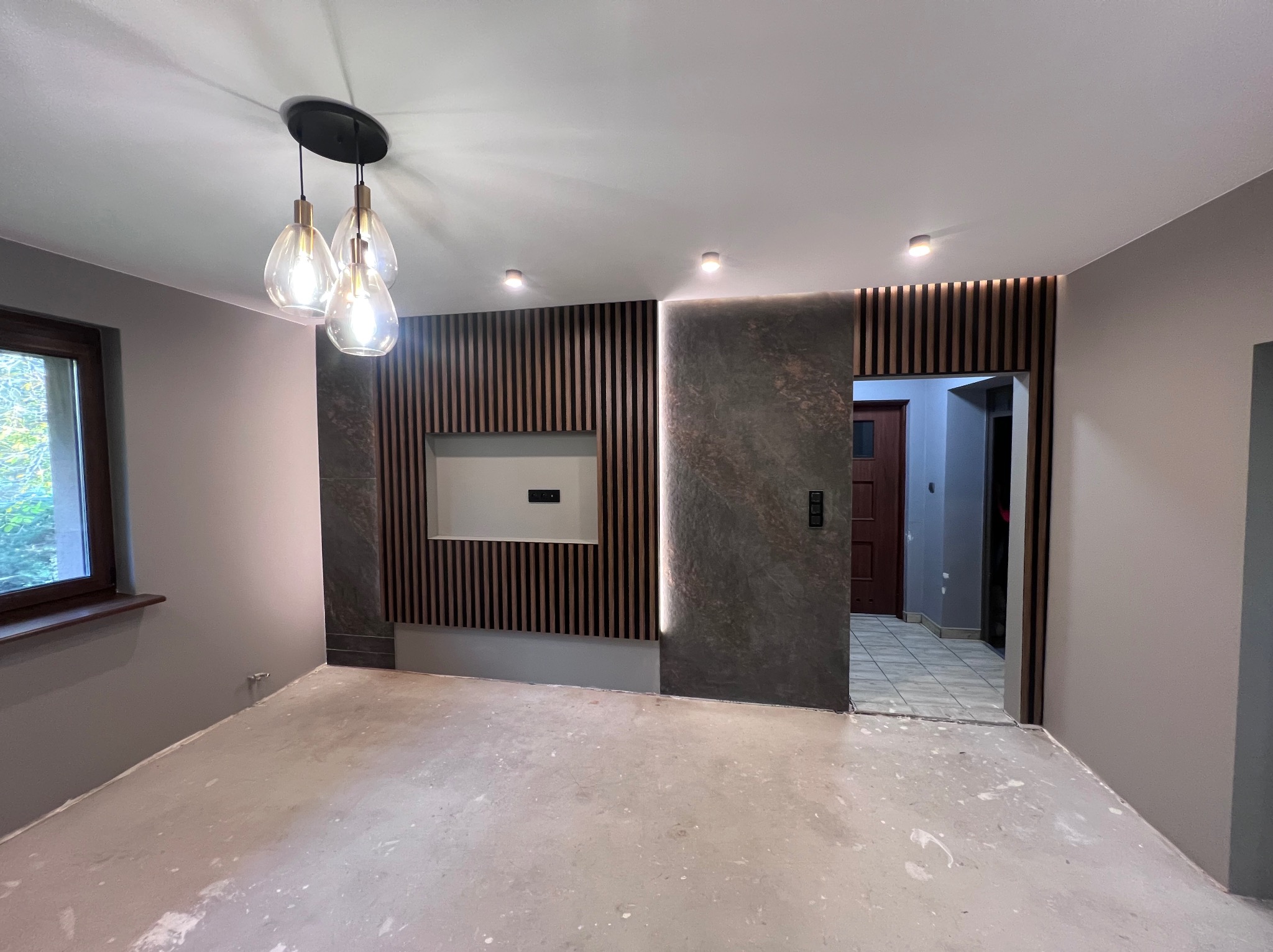 Nowoczesne wykończenie wnętrza: ściana z drewnianymi lamelami i kamienną płytą, designerska lampa wisząca, surowa podłoga przed położeniem paneli.