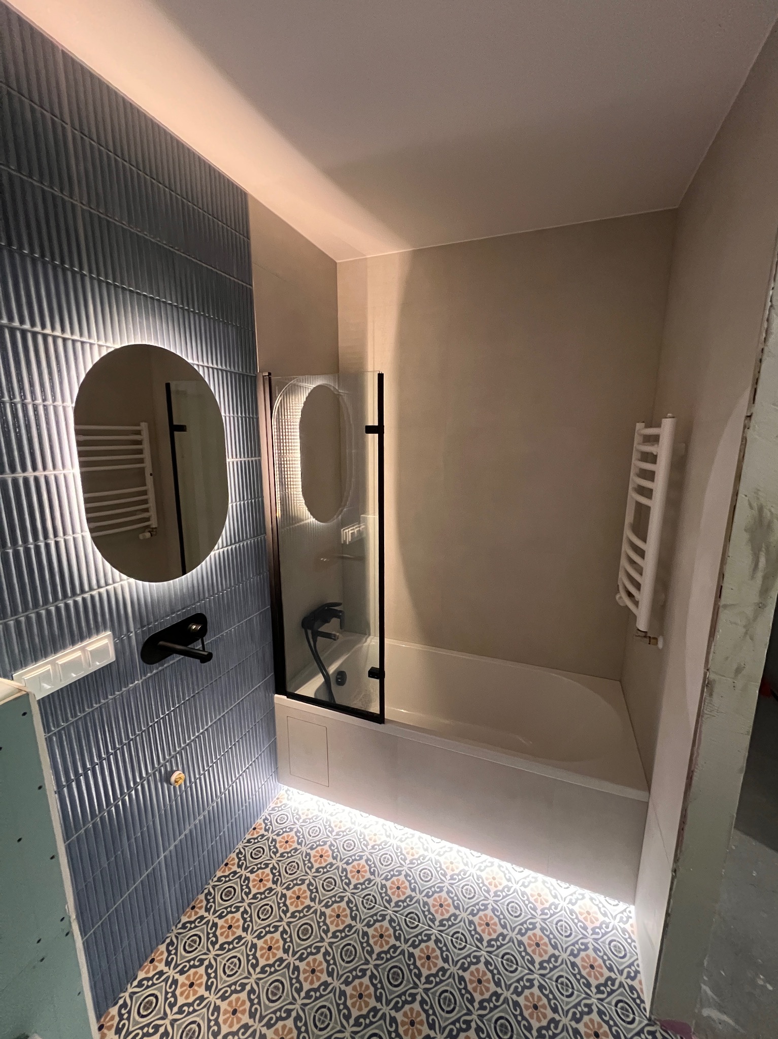 Wykończona łazienka z niebieskimi płytkami, okrągłym lustrem z podświetleniem LED, kabiną prysznicową i wzorzystą podłogą. Grzejnik drabinkowy na ścianie.