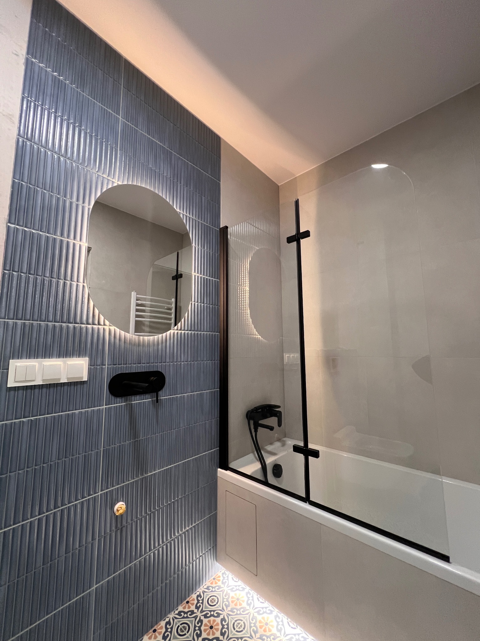 Nowoczesna łazienka z niebieskimi płytkami, okrągłym lustrem z podświetleniem i czarną armaturą. Na podłodze wzorzyste płytki. Kabina prysznicowa ze szklanym panelem.