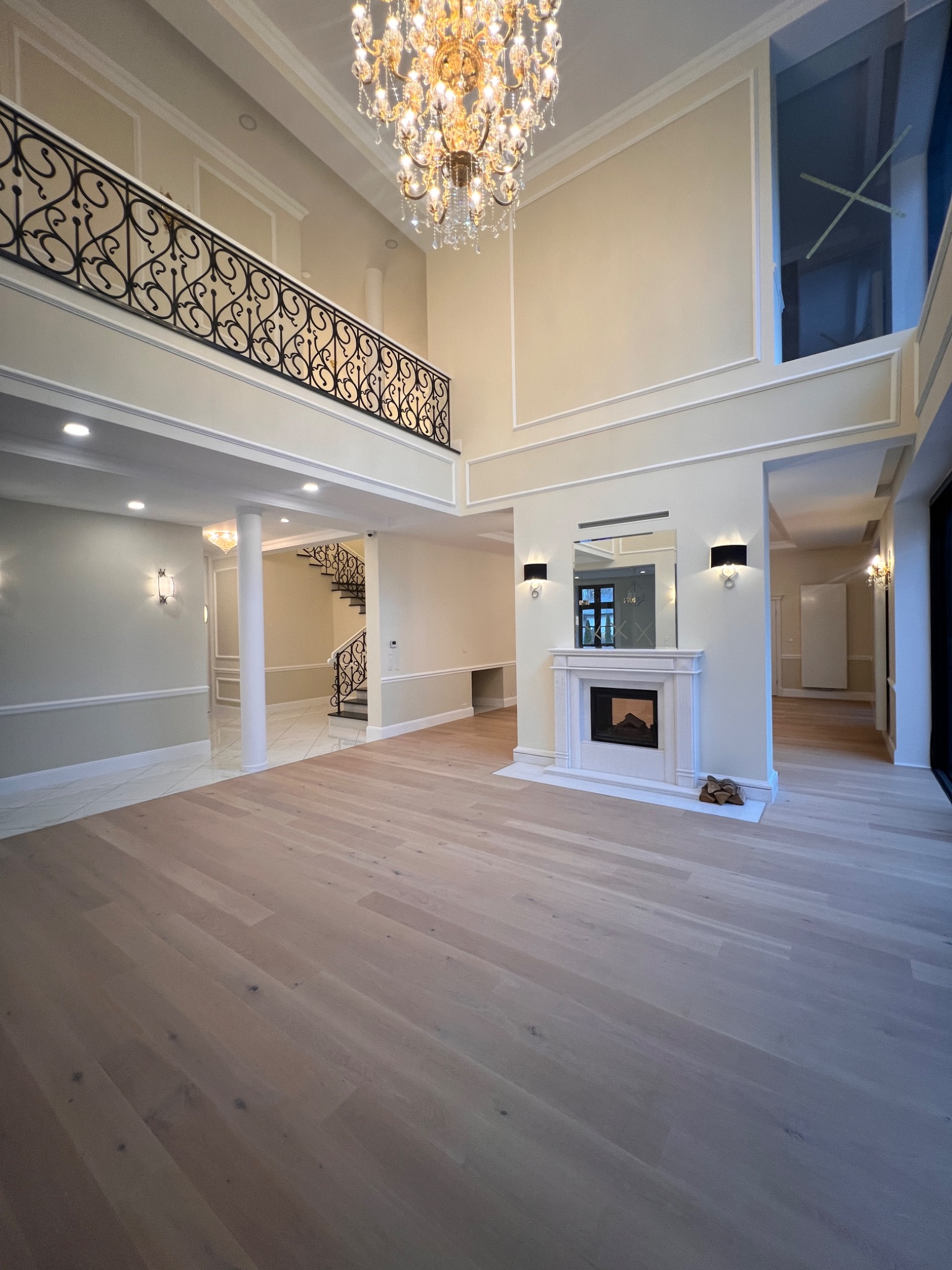 Przestronne wnętrze domu po remoncie: jasna podłoga, kremowe ściany z dekoracyjnymi listwami, żyrandol i balustrada z kutego żelaza. Widok na schody i kominek.