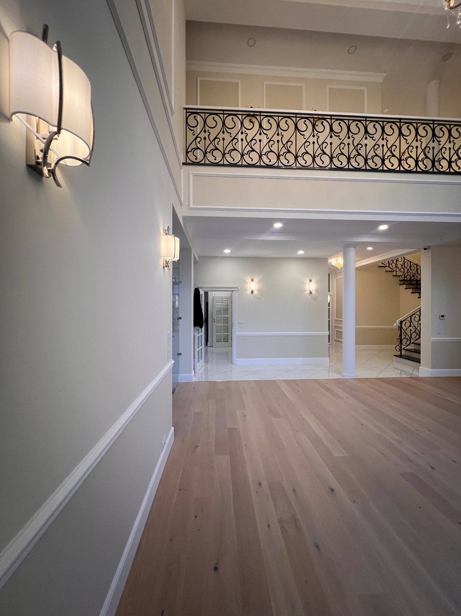 Eleganckie wnętrze domu z drewnianą podłogą, ozdobnymi listwami ściennymi, schodami z kutą balustradą i galerią na piętrze. Widoczne oświetlenie ścienne i marmurowe płytki w przejściu.