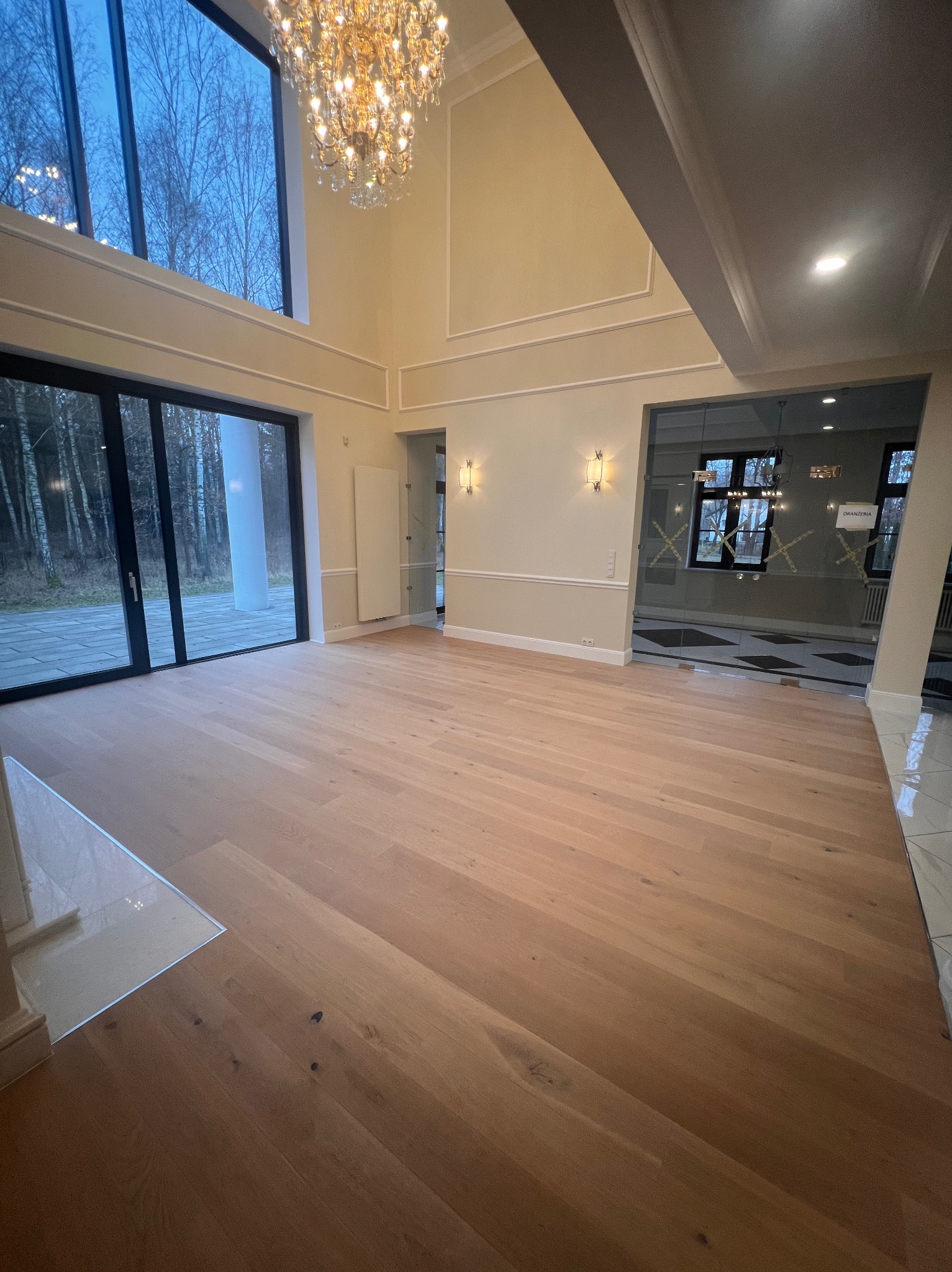 Jasna, drewniana podłoga w przestronnym wnętrzu z wysokimi oknami i ozdobnym żyrandolem. Widoczne detale wykończenia ścian i drzwi.