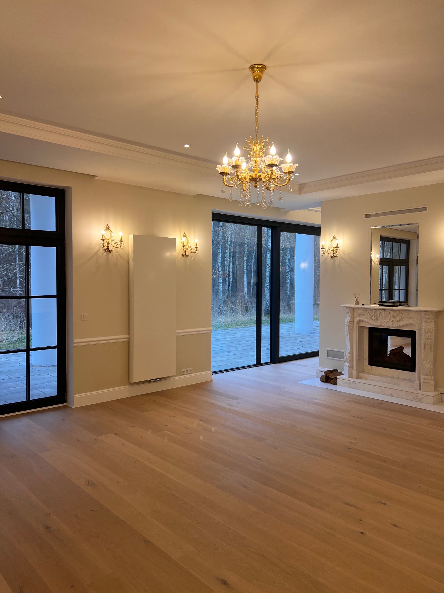 Jasne, przestronne wnętrze z parkietem, ozdobnym kominkiem i żyrandolem. Widoczne okna z czarnymi ramami i widok na zewnątrz. Eleganckie wykończenie.