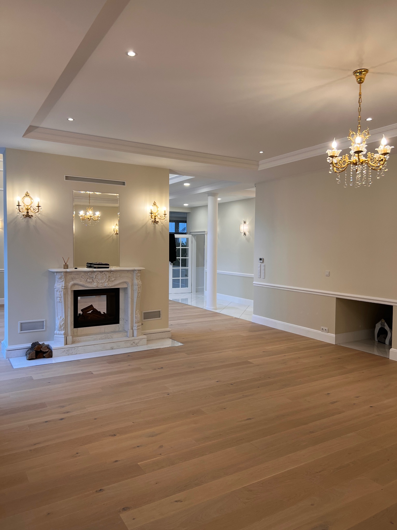 Eleganckie wnętrze z drewnianą podłogą, marmurowym kominkiem i kryształowym żyrandolem. Ściany w odcieniach beżu, widoczne detale sztukaterii i oświetlenia punktowego.