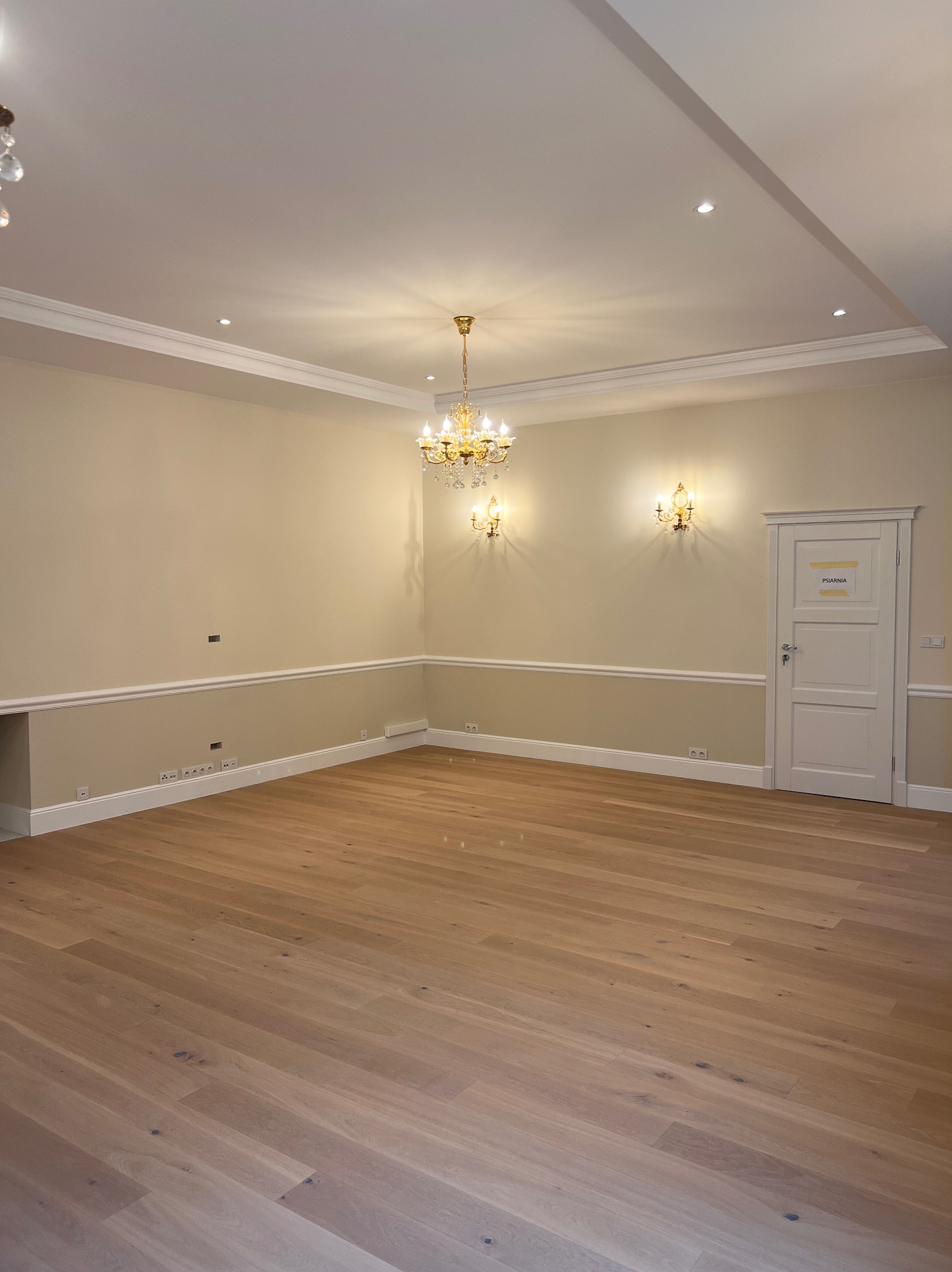 Jasne, przestronne wnętrze z drewnianą podłogą, kremowymi ścianami z listwami i ozdobnym żyrandolem. Widoczne gniazdka elektryczne i białe drzwi z napisem.
