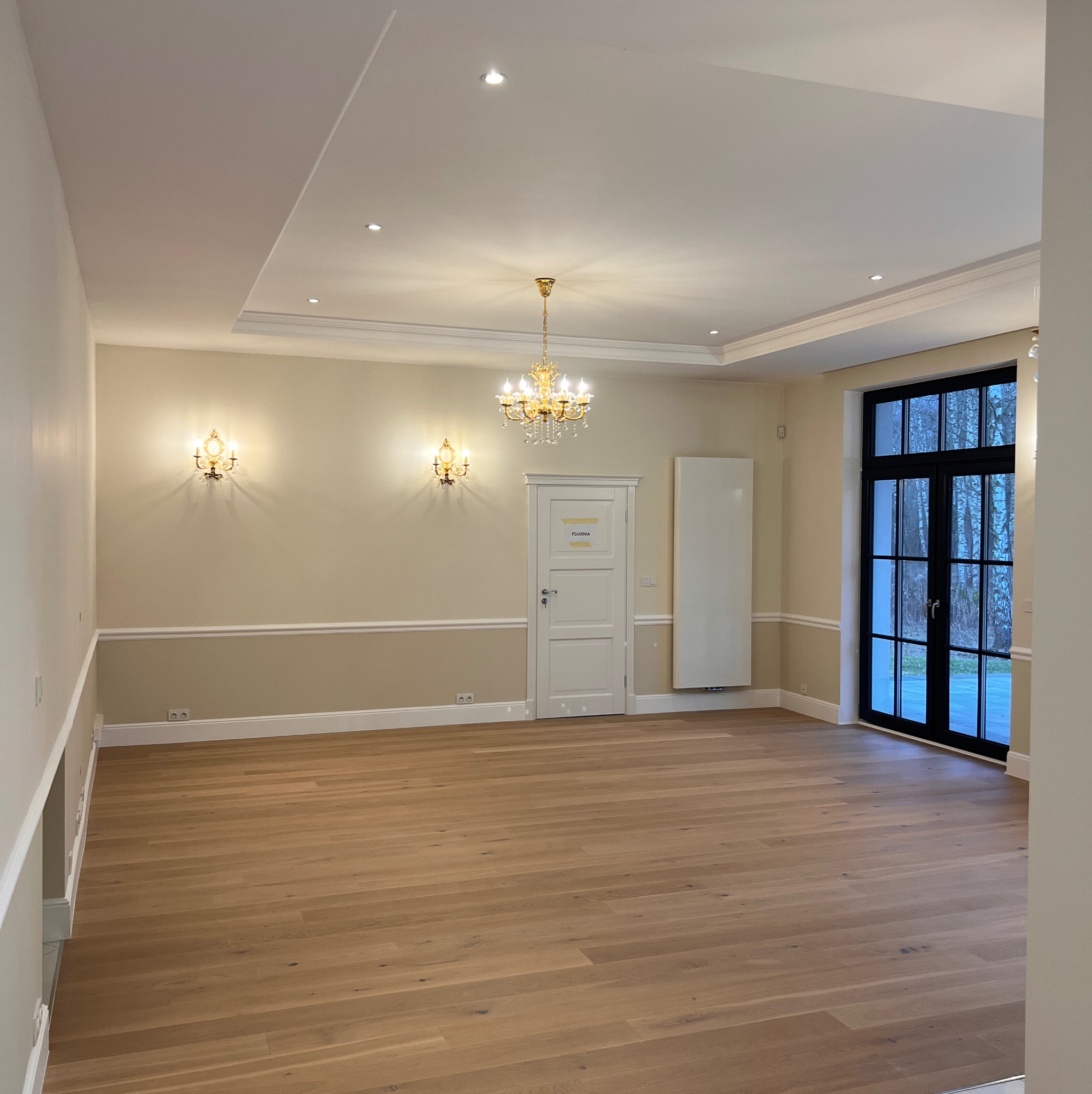 Eleganckie wnętrze po remoncie: parkiet, kremowe ściany z listwami, żyrandol i kinkiety. Drzwi z napisem 'Psiarnia'.