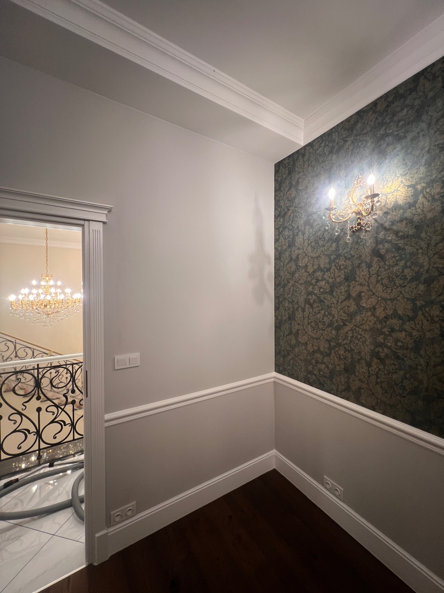 Eleganckie wykończenie wnętrza: ściana z wzorzystą tapetą i kinkietem, listwy przypodłogowe, widok na schody z kryształowym żyrandolem przez otwarte drzwi.