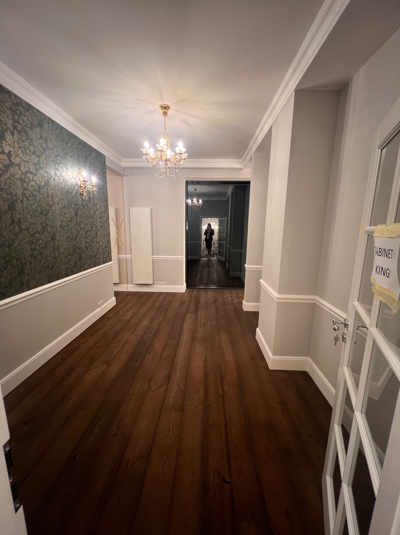 Korytarz po remoncie z ciemną drewnianą podłogą, szarą tapetą w kwiaty i kryształowym żyrandolem. Widoczne listwy przypodłogowe i drzwi z napisem.