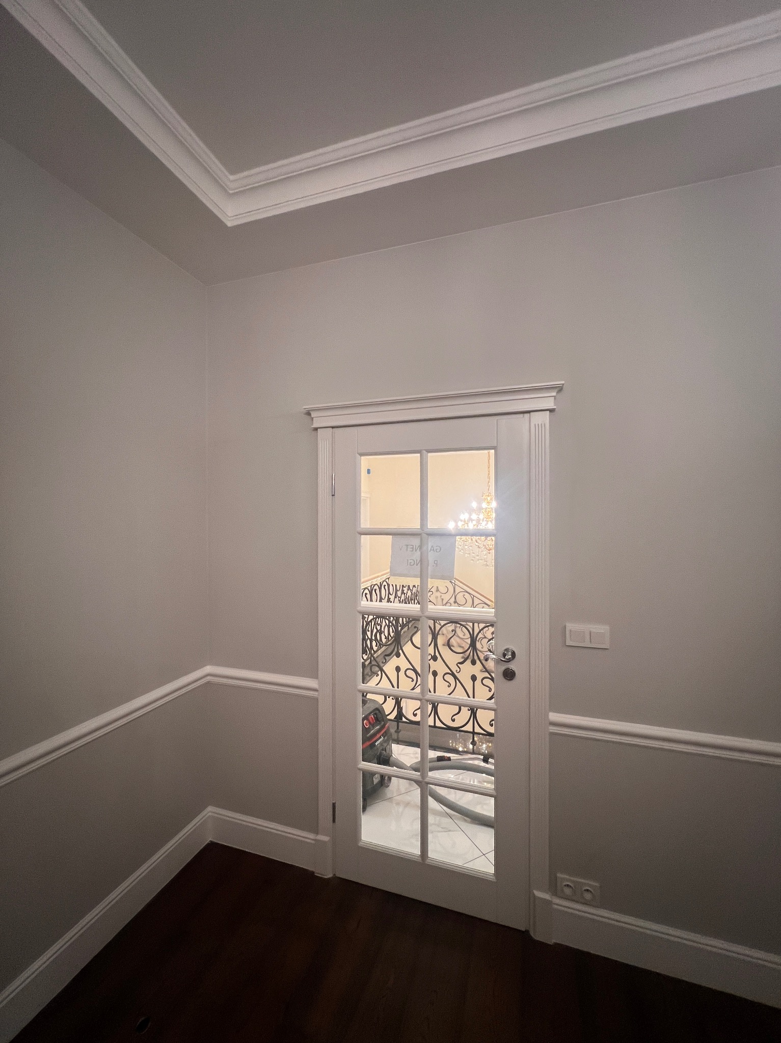 Eleganckie wnętrze: białe listwy przypodłogowe i sufitowe, szara ściana, drewniana podłoga, przeszklone drzwi z widokiem na schody z kutą balustradą i żyrandol.