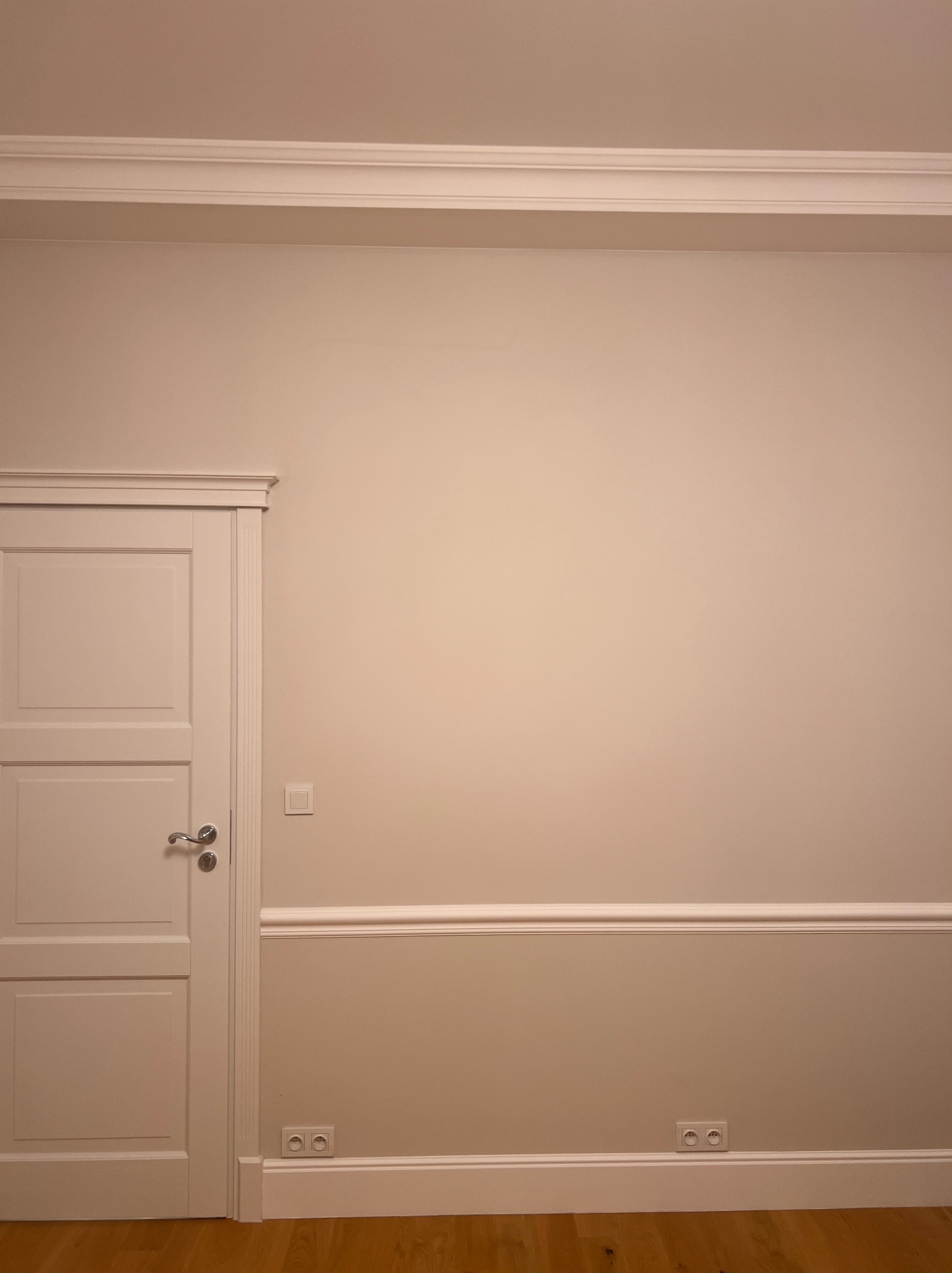 Jasne wnętrze po remoncie: kremowa ściana z dekoracyjną listwą, białe drzwi z ozdobną ramą, drewniana podłoga i gniazdka elektryczne. Minimalistyczny design.