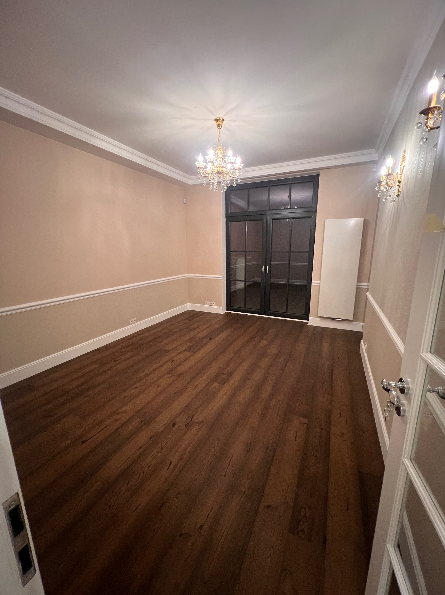 Eleganckie wnętrze po remoncie: parkiet w odcieniach brązu, kremowe ściany z białymi listwami, kryształowy żyrandol i przeszklone drzwi w czarnej ramie.