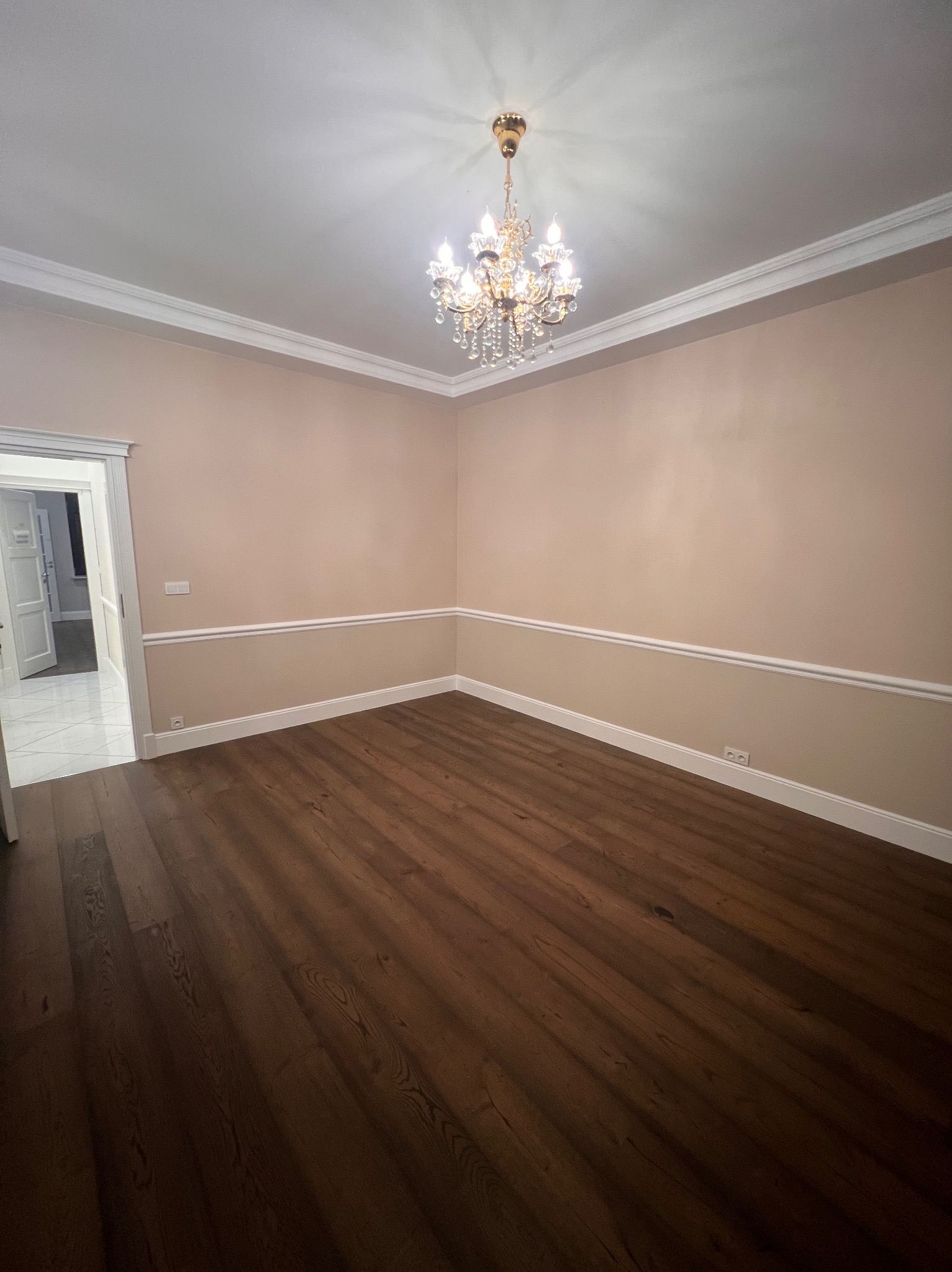 Pusty pokój po remoncie: ciemna podłoga, beżowe ściany z białymi listwami, kryształowy żyrandol. Widok na sąsiednie pomieszczenie z marmurową podłogą.
