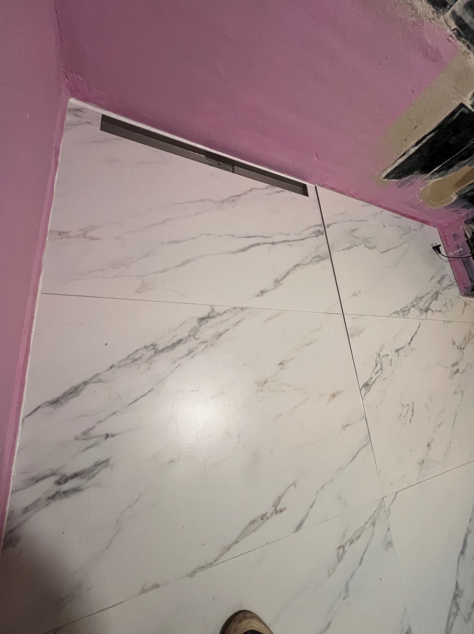 Ukończona podłoga w łazience z białych płytek imitujących marmur, z widocznym odpływem liniowym i fragmentem różowej ściany. Ujęcie z dołu.