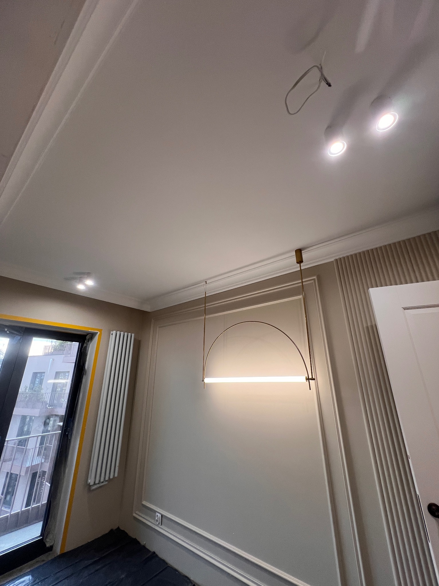 Wykończone wnętrze pokoju z nowoczesną lampą łukową, listwami dekoracyjnymi na ścianie i oknem z widokiem na budynek. Podłoga zabezpieczona folią.