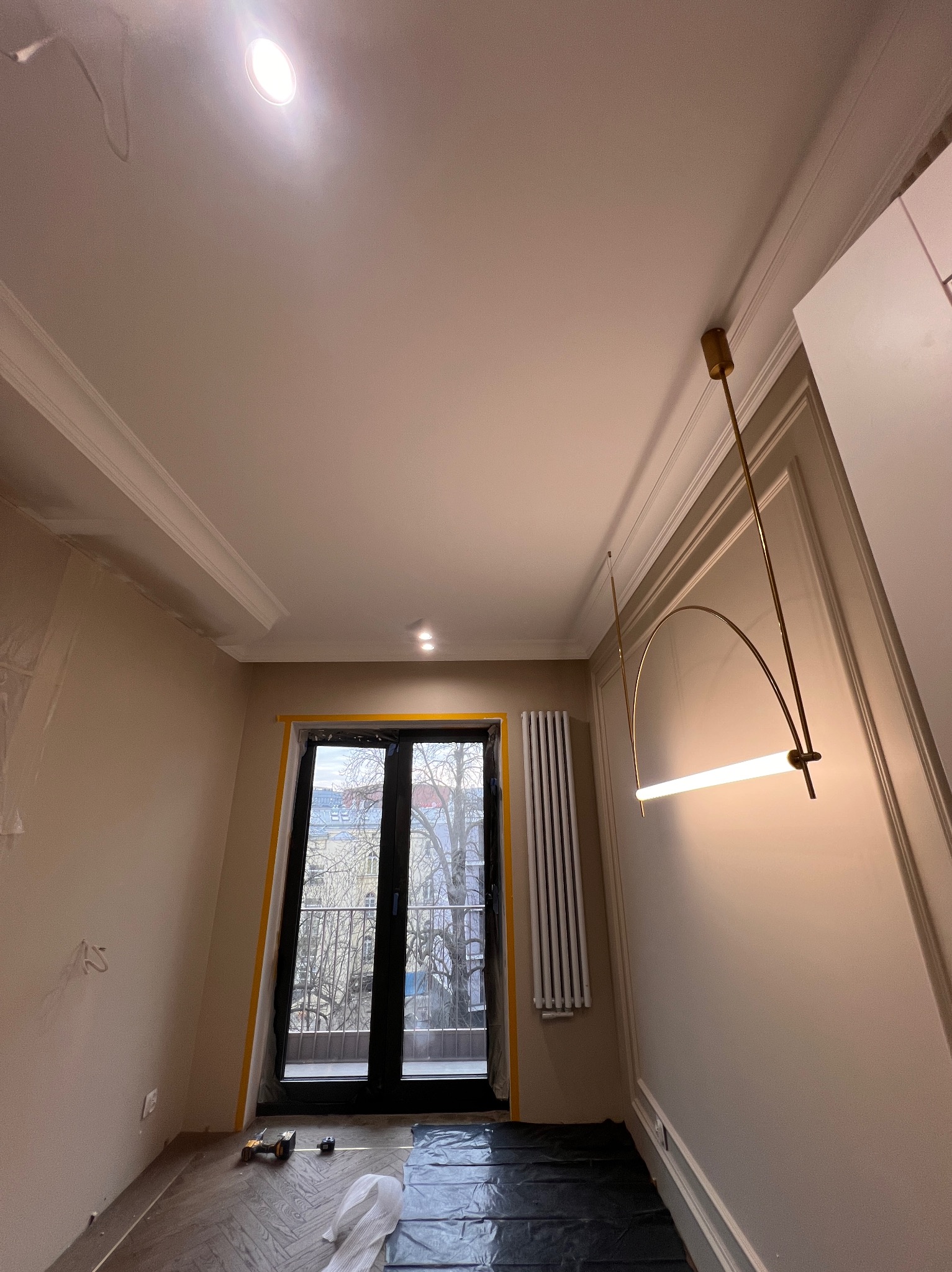 Wnętrze w trakcie wykańczania: parkiet w jodełkę, listwy przypodłogowe, grzejnik, okno balkonowe zabezpieczone taśmą, nowoczesna lampa. Widoczne narzędzia.