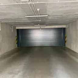 Zamykana brama garażowa w surowym, betonowym garażu podziemnym z oświetleniem. Widok na wprost, szara kolorystyka, minimalizm.