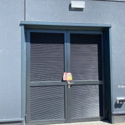 Tomasz Lipka Solid Doors - Dwuskrzydłowa, szara brama z żaluzjami na tle szarej ściany budynku, z zamontowanym znakiem ostrzegawczym na klamce. Widoczny fragment chodnika z kostki brukowej.