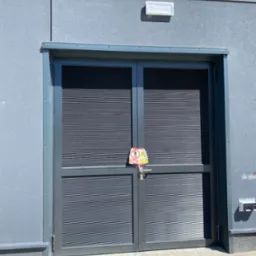 Dwuskrzydłowa, szara brama z żaluzjami na tle szarej ściany budynku, z zamontowanym znakiem ostrzegawczym na klamce. Widoczny fragment chodnika z kostki brukowej.