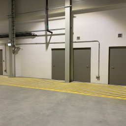 Tomasz Lipka Solid Doors - Trzy szare drzwi w hali przemysłowej z żółtymi pasami na podłodze. Widoczne instalacje pod sufitem i oświetlenie. Minimalistyczny design, surowe wykończenie.