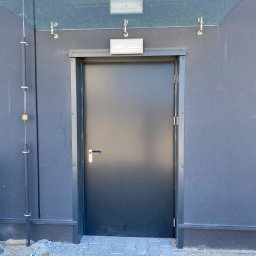 Tomasz Lipka Solid Doors - Zamknięte, grafitowe drzwi zewnętrzne z metalową ramą, umieszczone pod szklanym zadaszeniem na tle szarej ściany budynku. Widoczne elementy instalacji na ścianie.