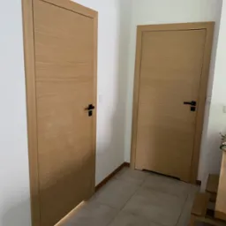 Dwa skrzydła drzwi wewnętrznych z naturalnego drewna, z czarnymi klamkami, zamontowane w jasnym korytarzu, z delikatnym podświetleniem dolnej krawędzi jednego skrzydła.