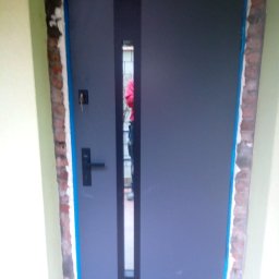 Tomasz Lipka Solid Doors - Szare drzwi zewnętrzne z pionowym przeszkleniem, widoczne ślady montażu i zabezpieczenia taśmą. Klucz w zamku, ceglana ościeżnica.