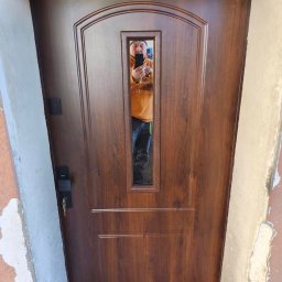 Tomasz Lipka Solid Doors - Brązowe drzwi zewnętrzne z szybą, widoczny zamek i klucz. W szybie odbija się osoba robiąca zdjęcie. Drzwi zamontowane w surowym otworze.