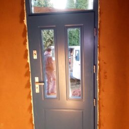 Tomasz Lipka Solid Doors - Niebieskie drzwi zewnętrzne z szybami, widoczne podczas montażu w surowym otworze. W szybach odbija się sylwetka osoby i zaparkowany biały samochód dostawczy.