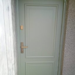 Tomasz Lipka Solid Doors - Zamknięte, jasne drzwi zewnętrzne z klamką i wizjerem, wkomponowane w szarą ścianę o ziarnistej fakturze. Widoczny fragment ościeżnicy i sąsiedniej ściany.