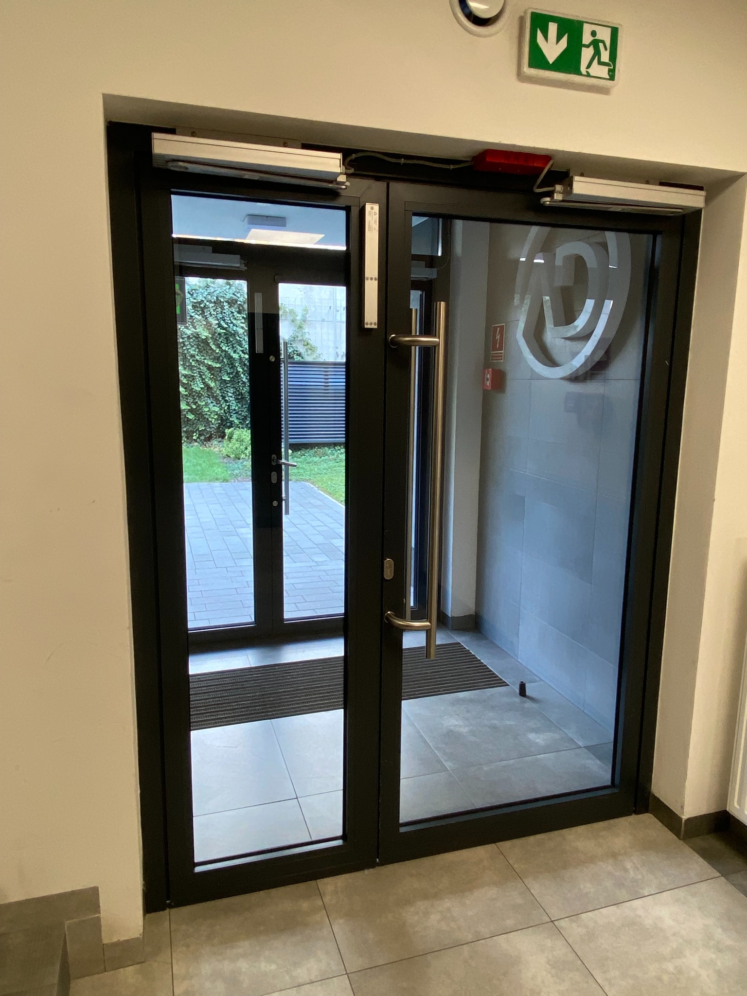 Szklane drzwi automatyczne z czarną ramą, widoczne zadaszenie i ogród na zewnątrz. Na ścianie znak ewakuacyjny i logo.
