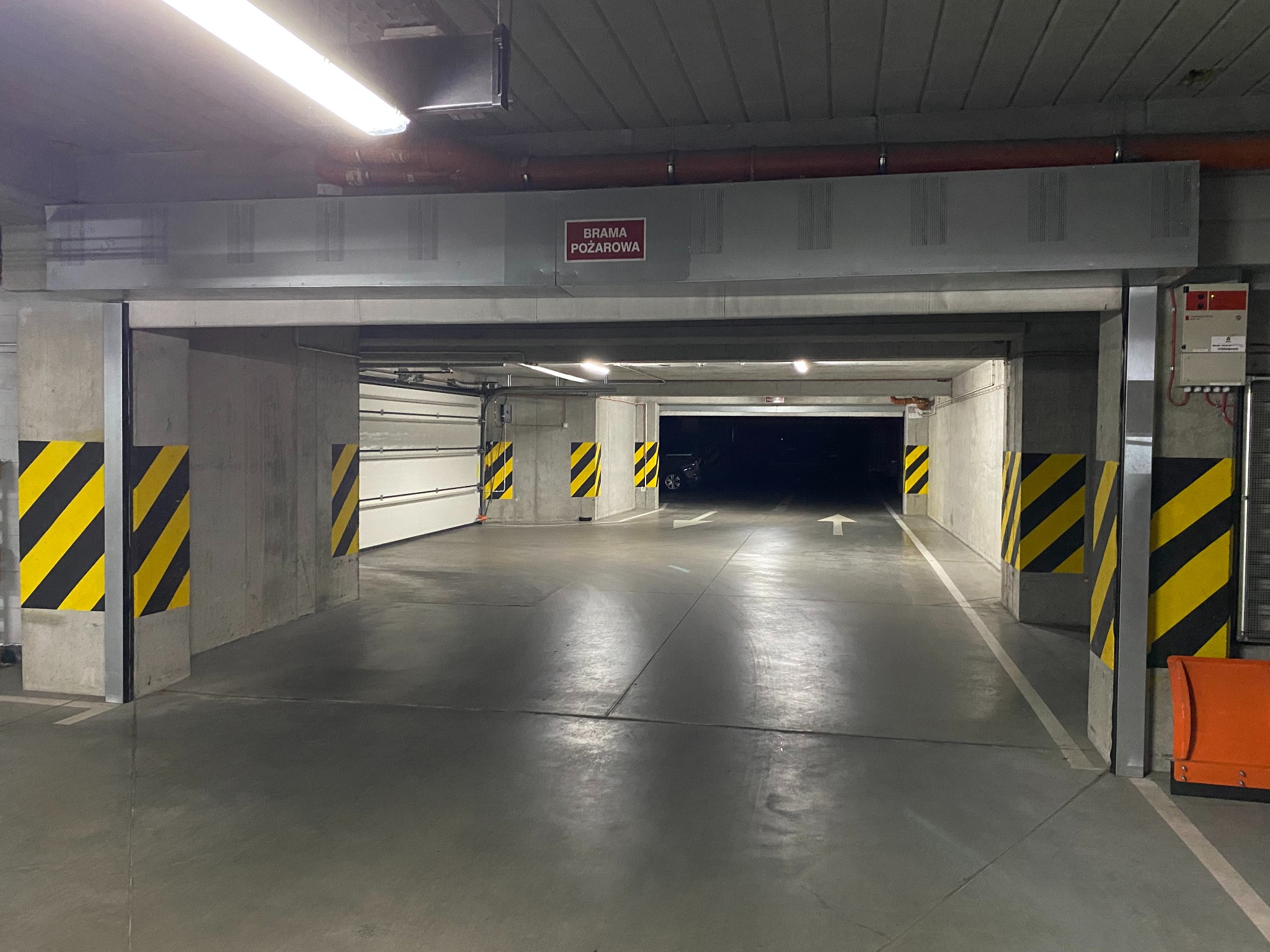 Wnętrze parkingu podziemnego z bramą przeciwpożarową, oznaczone słupy z żółto-czarnymi pasami, widoczny wjazd i zaparkowany samochód w oddali.