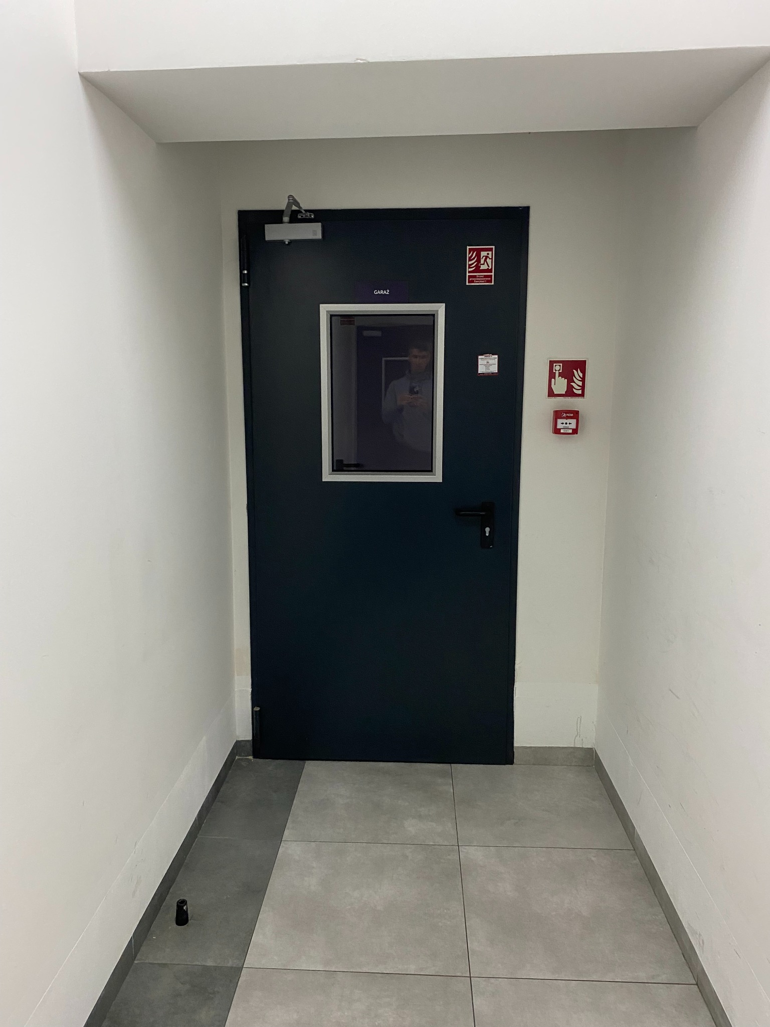 Ciemne drzwi techniczne z szybą w korytarzu, oznaczone jako 'Garaż', z widocznym systemem przeciwpożarowym na ścianie. Szara podłoga z płytek.