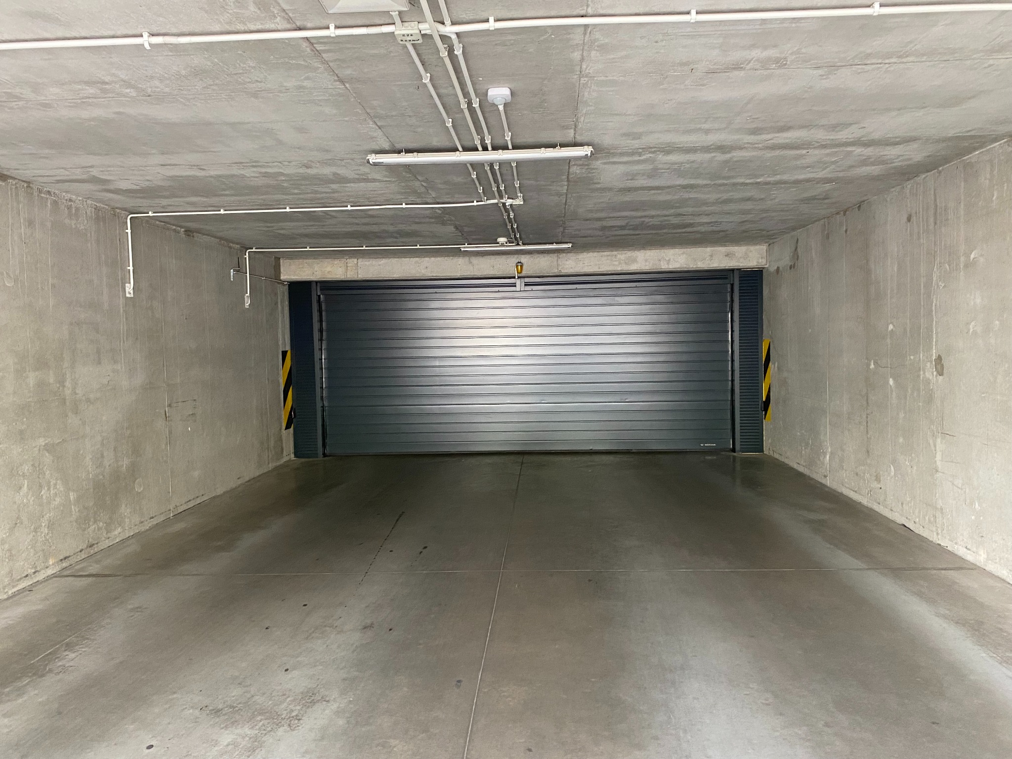 Zamykana brama garażowa w surowym, betonowym garażu podziemnym z oświetleniem. Widok na wprost, szara kolorystyka, minimalizm.