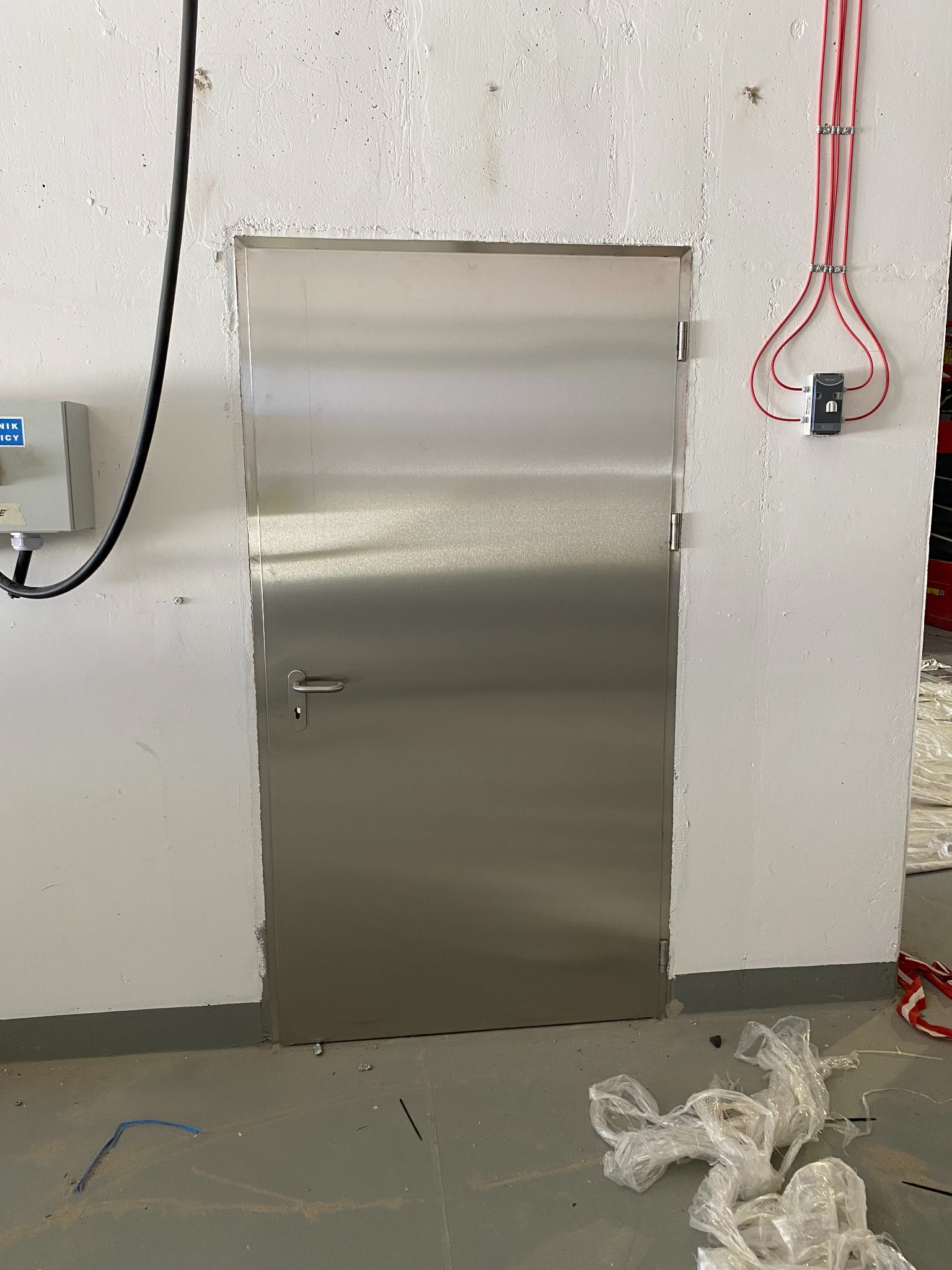 Metalowe drzwi techniczne w białej ścianie, widoczne przewody elektryczne i elementy instalacji. Prosty uchwyt, minimalistyczny design. Widok z bliska.