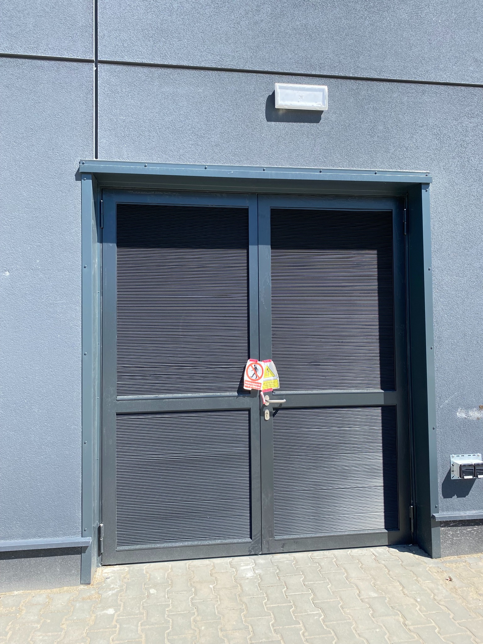 Dwuskrzydłowa, szara brama z żaluzjami na tle szarej ściany budynku, z zamontowanym znakiem ostrzegawczym na klamce. Widoczny fragment chodnika z kostki brukowej.