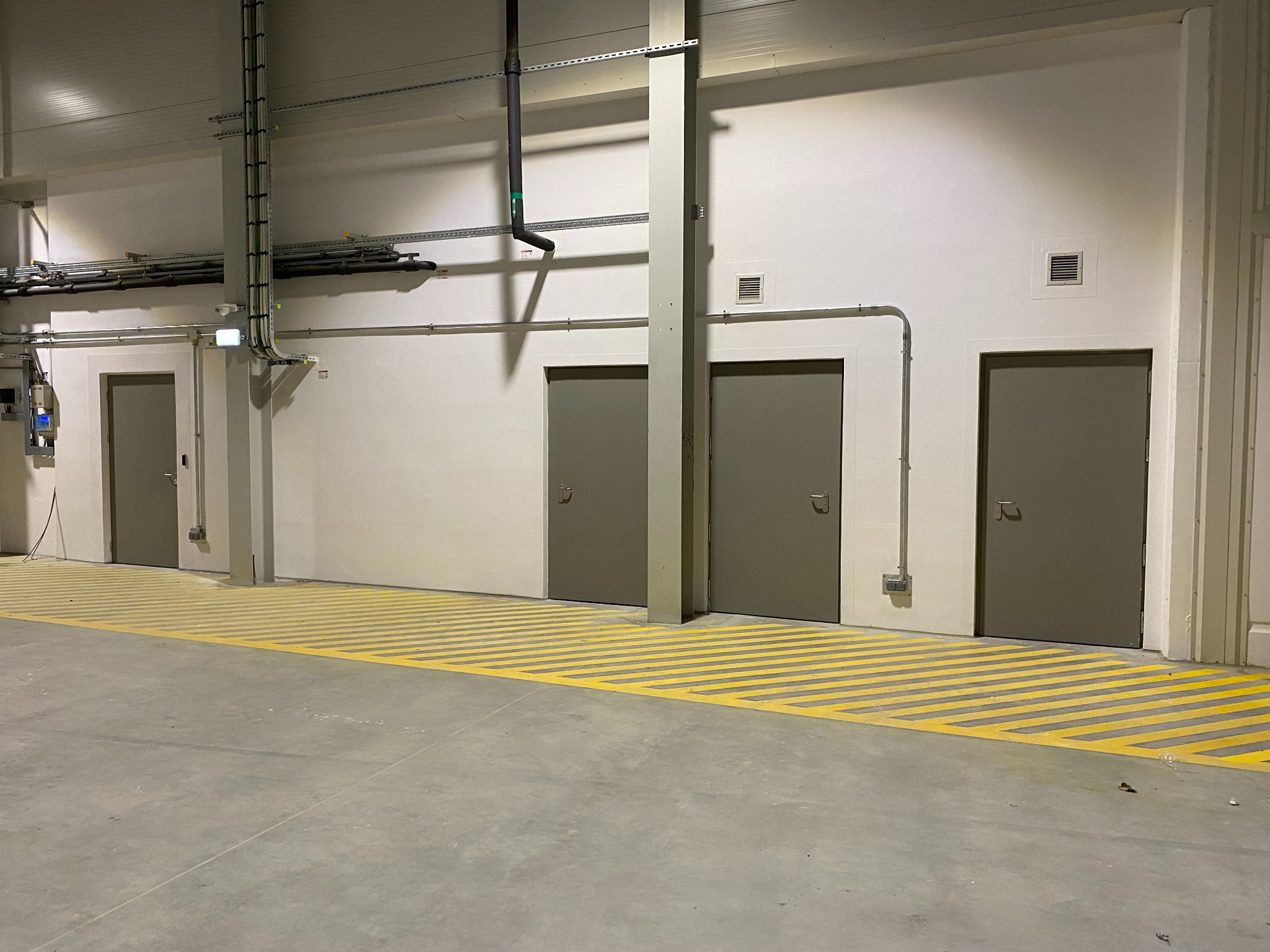 Trzy szare drzwi w hali przemysłowej z żółtymi pasami na podłodze. Widoczne instalacje pod sufitem i oświetlenie. Minimalistyczny design, surowe wykończenie.