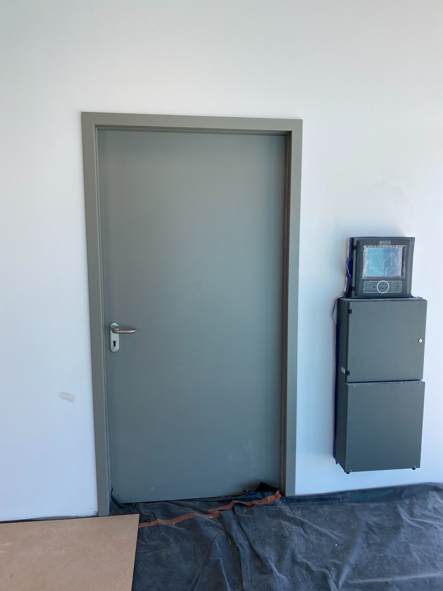 Szare, metalowe drzwi z klamką w białej ścianie. Obok zamontowana skrzynka sterownicza. Podłoga zabezpieczona folią malarską.