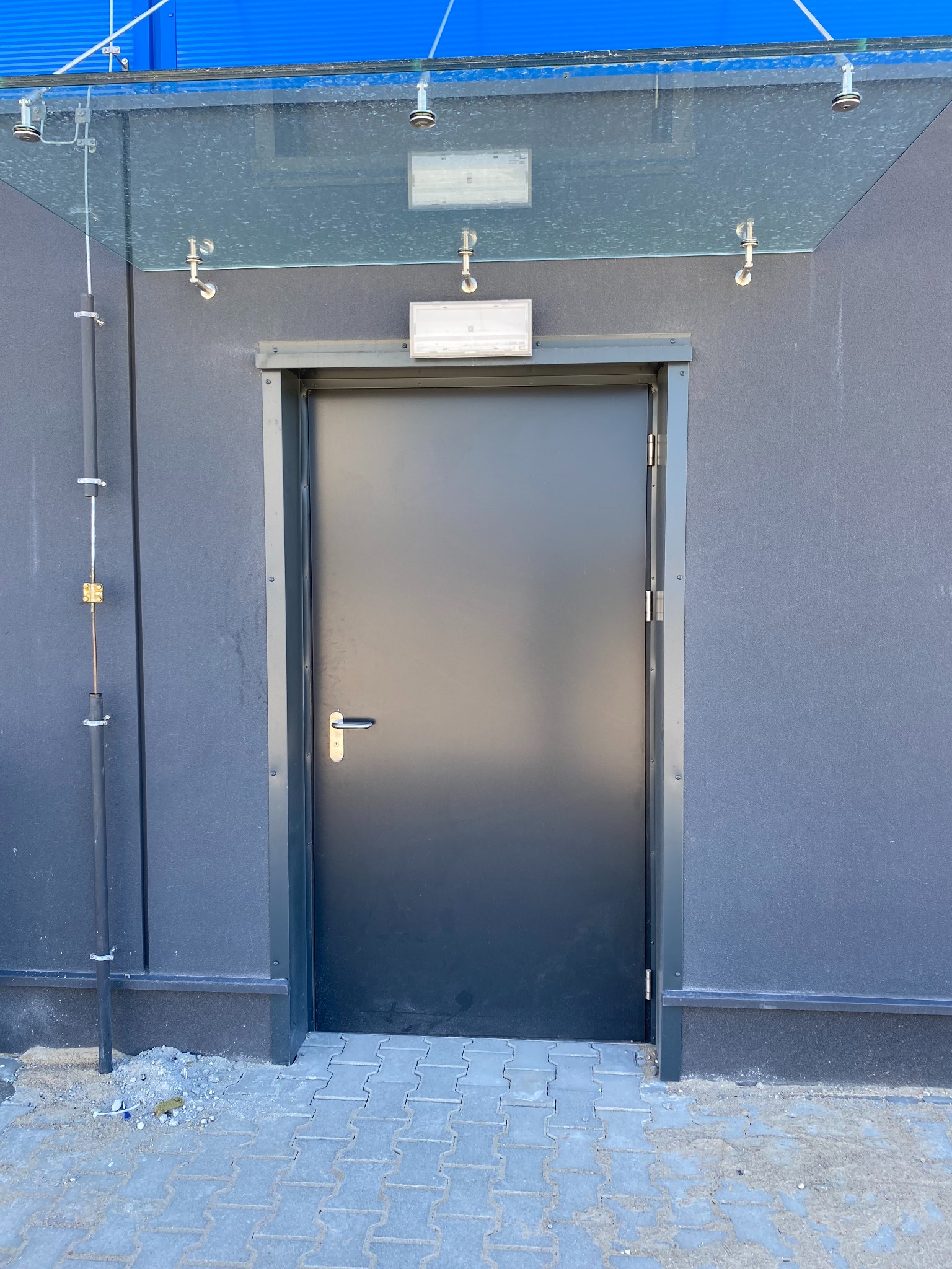 Zamknięte, grafitowe drzwi zewnętrzne z metalową ramą, umieszczone pod szklanym zadaszeniem na tle szarej ściany budynku. Widoczne elementy instalacji na ścianie.